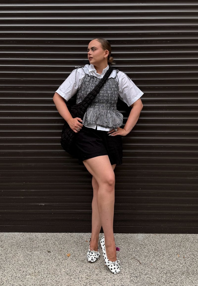 Young woman wearing a white blouse with a checkered top, black shorts, polka dot shoes, and a black shoulder bag, standing against a dark wall.