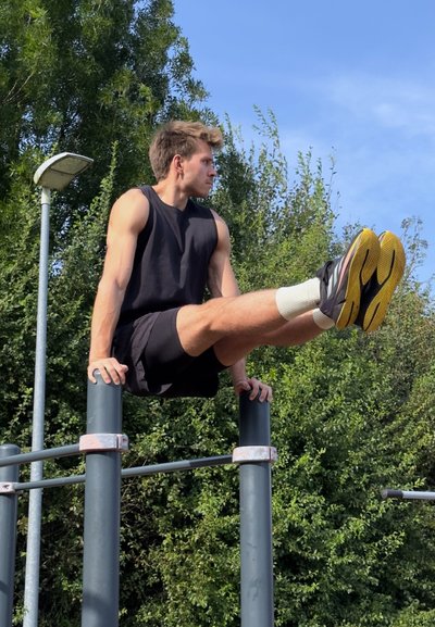 Un joven con una camiseta sin mangas negra y pantalones cortos realiza un ejercicio de calistenia en barras paralelas frente a un fondo verde.