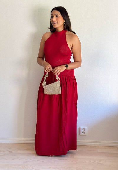 Top rojo con cuello halter y una larga falda fluida. La modelo sostiene un pequeño bolso metálico dorado. Textura suave, corsé ajustado y falda de pliegues.