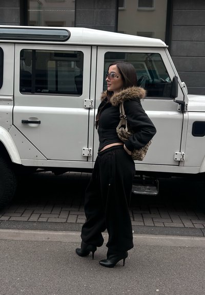 Mujer con cabello largo y oscuro, vistiendo chaqueta con cuello de piel negra, bolso, pantalones sueltos y botas de tacón, de pie junto a un vehículo todoterreno blanco en una calle de la ciudad.