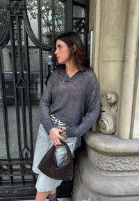 Šedý pletený svetr s výstřihem do V, stříbrné kožené šortky, černá pletená kabelka a pruhovaný pásek. Stojí poblíž zdobených kovaných dveří.