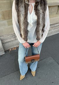 Colete de pelo castanho sobre blusa branca com babados, combinado com jeans de perna larga azul claro. Bolsa castanha e estampada, sapatos bege de bico fino.