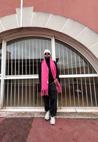 Άνδρας που φοράει ένα ροζ κασκόλ και σκουφάκι, μαύρη ζακέτα και χαλαρούς μαύρους παντελόνες, στέκεται μπροστά από μια γυάλινη πόρτα με κάθετους στύλους.