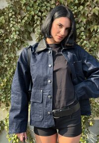 Dunkelblaue Jeansjacke mit schwarzem Lederkragen, silbernen Druckknöpfen und Fronttaschen, getragen über einem schwarzen T-Shirt und Shorts.