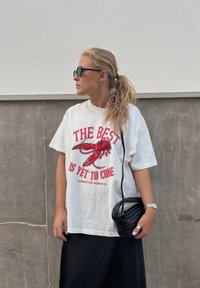 Valge puuvillane t-särk punase homaarigraafika ja tekstiga, kombineeritud musta pika seeliku ja väikesega punutud musta kottiga.