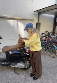 Donna con felpa gialla e cappellino blu che sistema una borsa marrone su uno scooter Vespa nero accanto a biciclette parcheggiate.