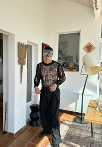 Ung man med tatueringar som bär en svart nättröja och keps står inomhus nära en skulptur och en golvlampa, med solljus på kakelgolvet.