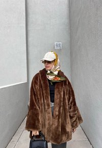 Καφέ ψεύτικη γούνα oversized παλτό με φαρδιές μανίκες, συνδυασμένο με μαύρη μπλούζα και γκρι παντελόνι. Διακοσμημένο με ένα μοτίβο μεταξωτό κασκόλ και καπέλο.