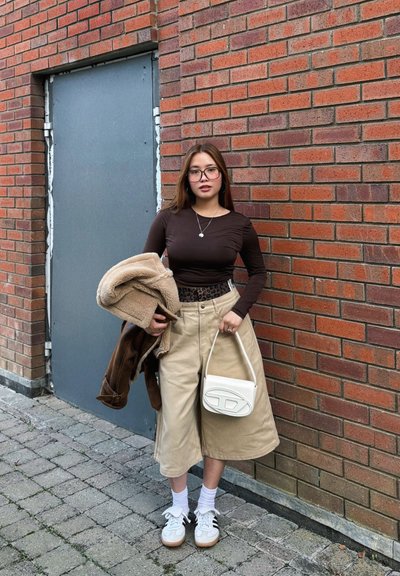 Blusa marrón de manga larga, pantalones anchos beiges, bolso blanco y zapatillas blancas. Los accesorios incluyen un collar y un abrigo marrón. Fondo de ladrillo.