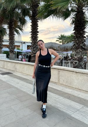 Un vestido maxi negro con un corsé ajustado y una falda acampanada, combinado con un cinturón decorativo. Zapatillas negras con acentos blancos y un bolso plateado.