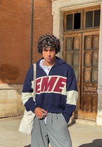 Μπλε ναυτικού φούτερ με φερμουάρ και έντονη επιγραφή "EME" σε μπορντό, κρεμ μανίκια, συνδυασμένο με ανοιχτού γκρι πλισέ παντελόνι και τσάντα ώμου.