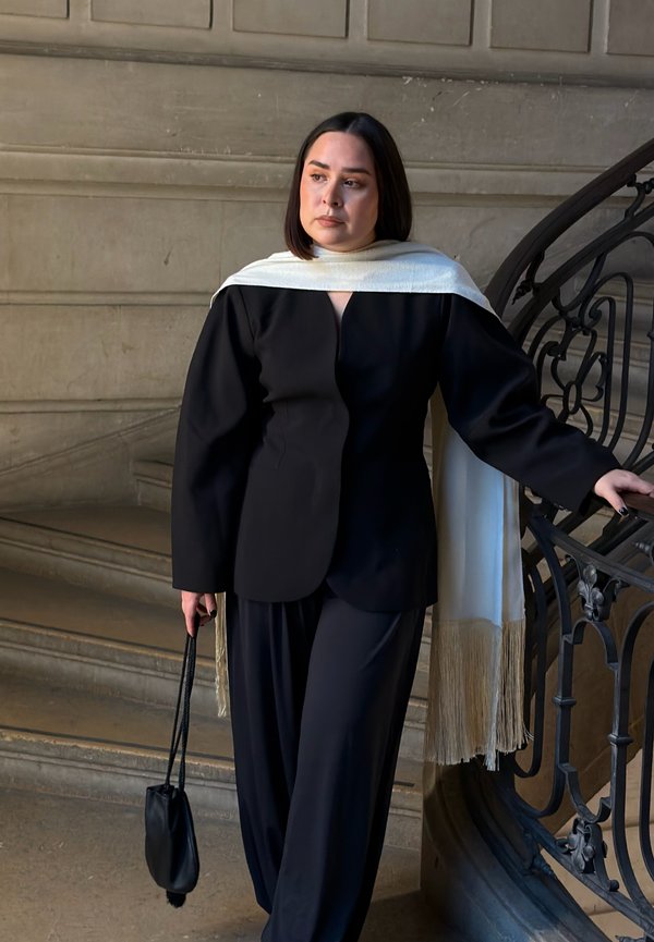 Une femme en tenue noire avec un châle à franges blanches s'appuie sur la rampe ornée d'un escalier, tenant un petit sac à main noir, debout sur des marches en pierre.