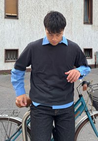 Felpa nera a girocollo indossata sopra una camicia azzurro chiaro con bottoni, abbinata a jeans scuri. La persona è in piedi accanto a una bicicletta.