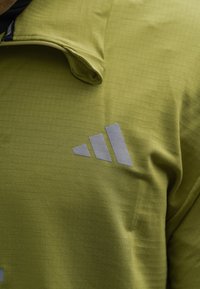 Grön kortärmad skjorta med vikta kragekanter och tre grå diagonala staplar som logotyp på bröstet.