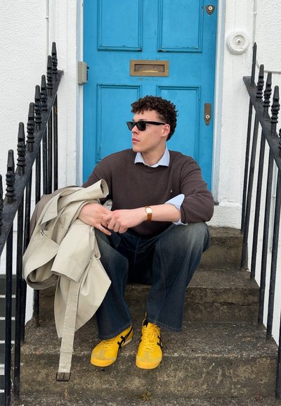 Suéter marrón sobre una camisa azul claro, jeans oscuros de pierna ancha, zapatillas amarillas con detalles en negro, y un abrigo beige colgado del brazo.