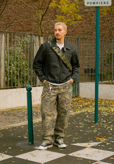 Chaqueta vaquera con bolsillos parche, pantalones de camuflaje y zapatillas negras. Incluye una correa verde para el hombro y accesorios de llavero.