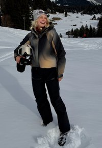 Χαμογελαστή γυναίκα με εξοπλισμό σκι κρατώντας κράνος στέκεται σε βαθύ χιόνι με χιονισμένο ορεινό χωριό και δέντρα στο φόντο.