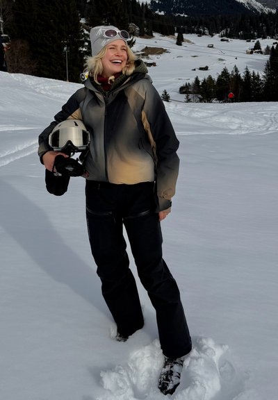 Mujer sonriente con equipo de esquí sosteniendo un casco, de pie en nieve profunda con un pueblo montañoso nevado y árboles en el fondo.