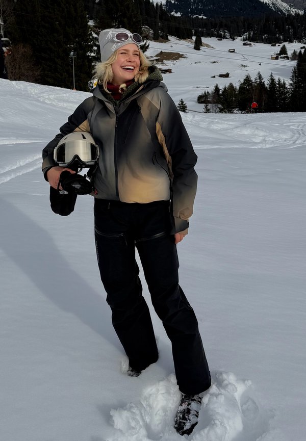 Χαμογελαστή γυναίκα με εξοπλισμό σκι κρατώντας κράνος στέκεται σε βαθύ χιόνι με χιονισμένο ορεινό χωριό και δέντρα στο φόντο.