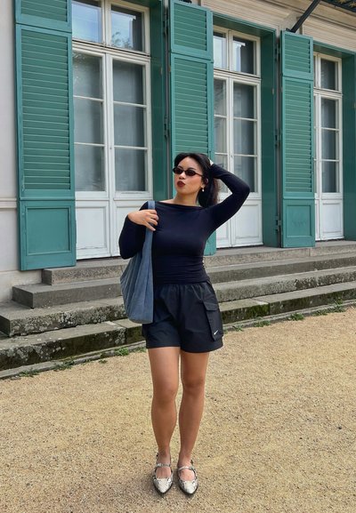 Top de manga larga negro, pantalones cortos negros con bolsillos, bolsa de mano gris y zapatos plateados. Contraste con un fondo de contraventanas turquesas y escalones.