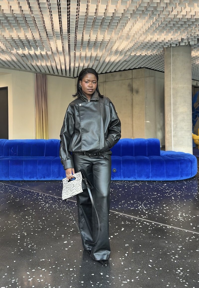 Femme en veste et pantalon en cuir noir tenant un sac à main blanc à motifs, debout devant un canapé en velours bleu dans un hall moderne.