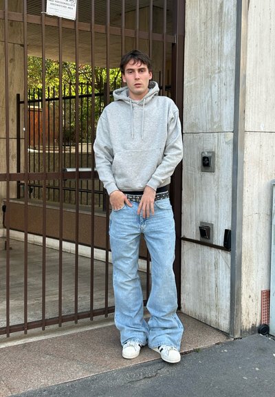 Joven con sudadera gris y vaqueros anchos azul claro de pie junto a una puerta metálica cerrada adherida a la pared de un edificio de piedra.