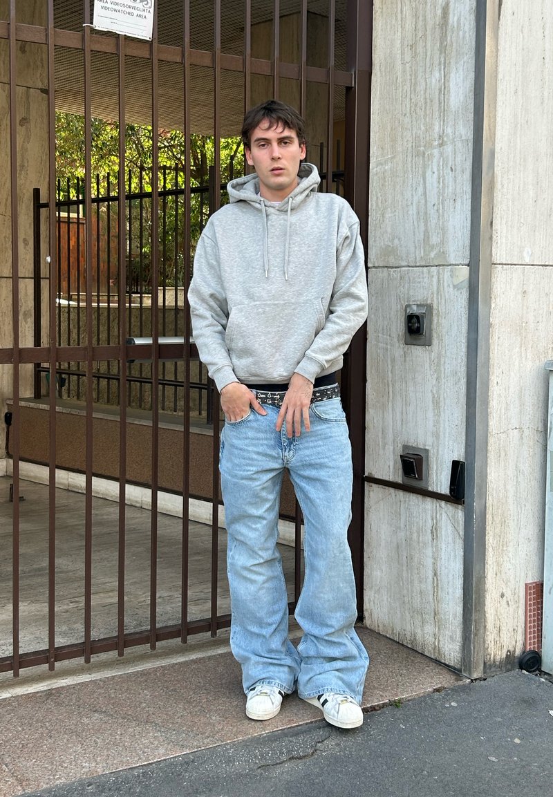 Mladý muž v sivom kapucniaku a voľných svetlomodrých džínsoch stojí pri zatvorenej kovovej bráne pripevnenej k kamennej stene budovy.