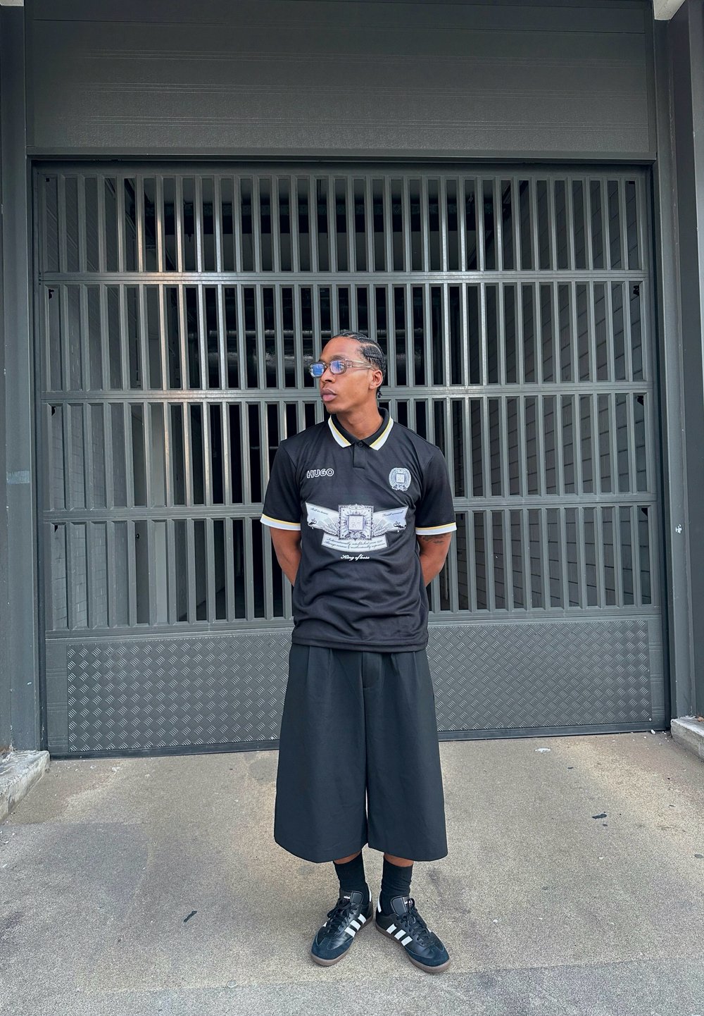 Μαύρη μπλούζα κοντό μανίκι με σχέδιο στο μπροστινό μέρος, κίτρινες λεπτομέρειες, συνδυασμένη με φαρδιά μαύρα σορτς και μαύρα αθλητικά παπούτσια. Στηρίζεται σε μια μεταλλική πύλη.