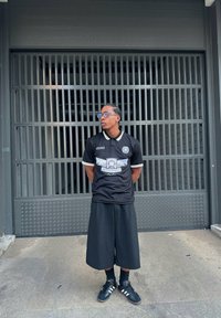 Μαύρη μπλούζα κοντό μανίκι με σχέδιο στο μπροστινό μέρος, κίτρινες λεπτομέρειες, συνδυασμένη με φαρδιά μαύρα σορτς και μαύρα αθλητικά παπούτσια. Στηρίζεται σε μια μεταλλική πύλη.