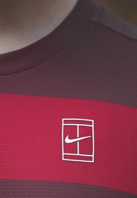 Raudona ir tamsiai raudona Nike sportinė marškinė su tekstūruotu raštu ir baltu logotipu ant krūtinės. Trumpomis rankovėmis ir pritaikyta stiliumi.