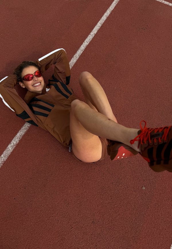 Γυναίκα με καφέ αθλητικά ρούχα και κόκκινα γυαλιά ηλίου ξαπλωμένη σε κόκκινο στίβο, χαμογελώντας με τα πόδια σηκωμένα και σταυρωμένα, εμφανείς λευκές γραμμές διαδρόμων.