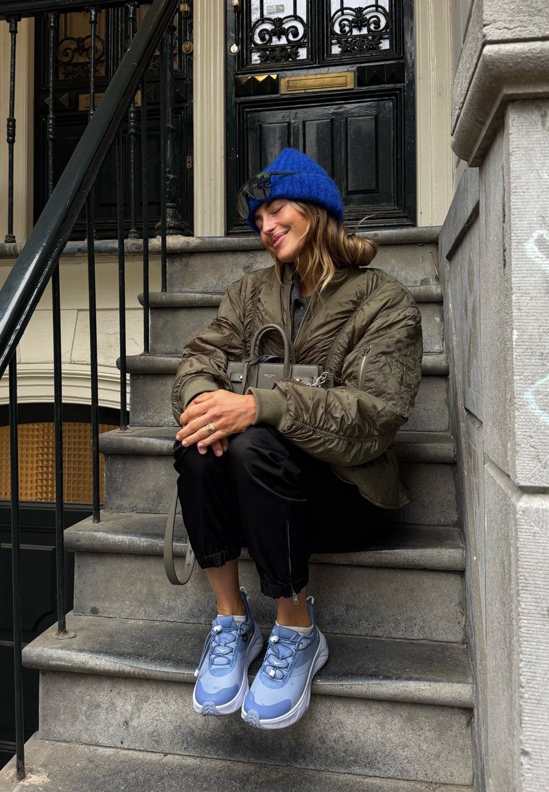 Kék sapkát viselő nő, olívazöld kabátban és világoskék tornacipőben, kőlépcsőn ül fekete ajtó előtt, szürke kézitáskát tartva.