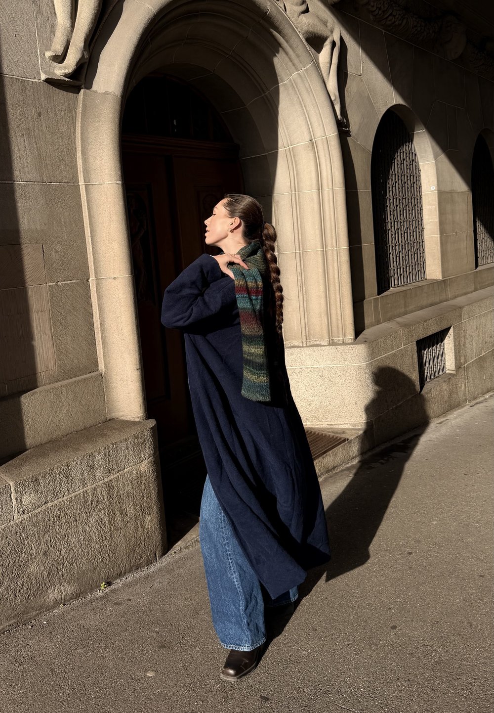 Gara tumši zila mētelis pār zilām zvana biksēm, kopā ar daudzkrāsainu adītu šalli. Fons ir ar akmens arku un ēnas detaļām.