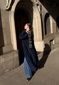 Gara tumši zila mētelis pār zilām zvana biksēm, kopā ar daudzkrāsainu adītu šalli. Fons ir ar akmens arku un ēnas detaļām.