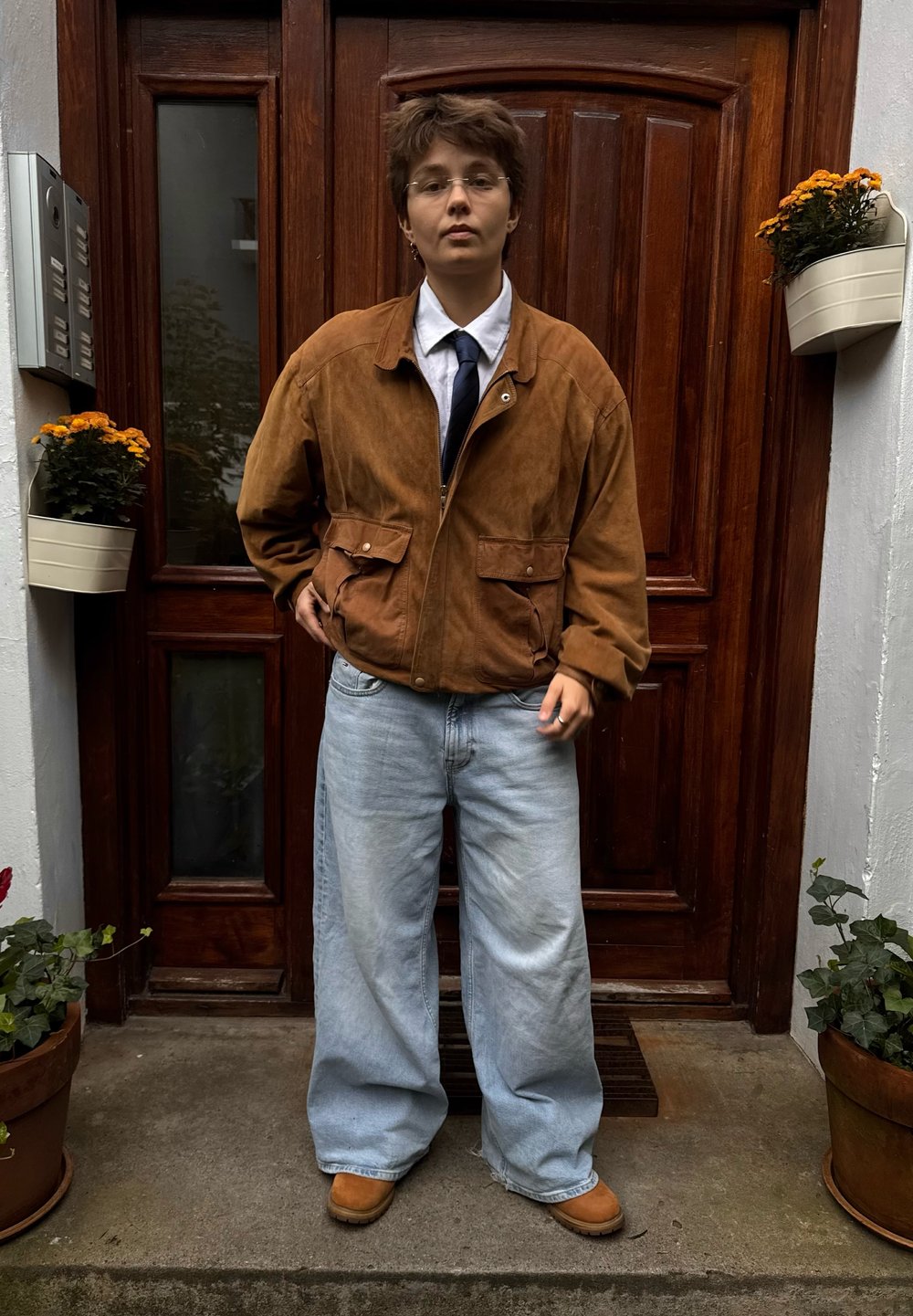 Hnedá semišová bunda, biela košeľa, námornícka kravata a voľné svetlomodré džínsy. Hnedé čižmy dotvárajú outfit. V pozadí sú drevené dvere a kvety v kvetináčoch.