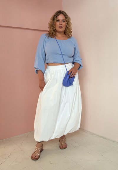 Top corto texturizado de color azul claro; falda larga y fluida blanca; bolso pequeño cruzado morado; sandalias marrones con tiras; fondo neutro.