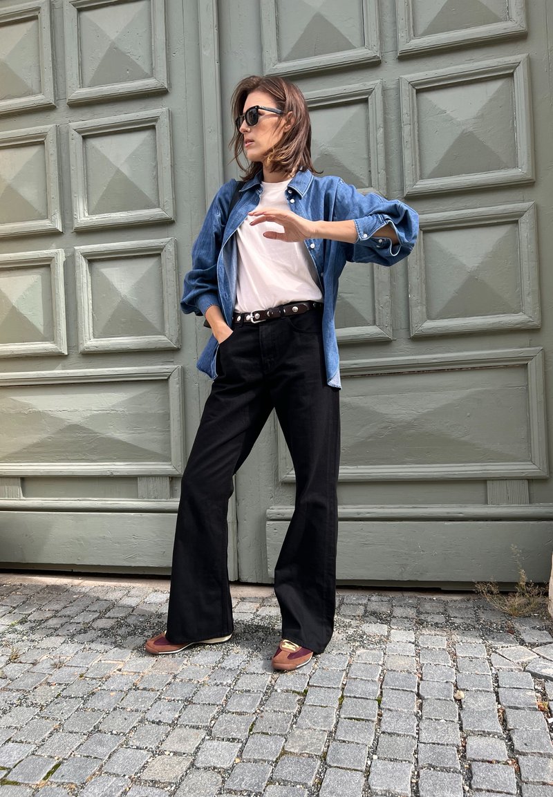 Τζιν πουκάμισο, λευκό μπλουζάκι, μαύρο παντελόνι με φαρδιά πόδια, καφέ και μπορντώ παπούτσια. Στηρίζεται σε μια υφασμάτινη πράσινη τοιχογραφία, με τα χέρια στη μέση.