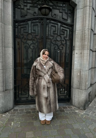 Abrigo de piel sintética en tonos marrones claros, con diseño ceñido, llevado sobre una blusa blanca, combinado con pantalones blancos y botas marrones, frente a una puerta decorativa.