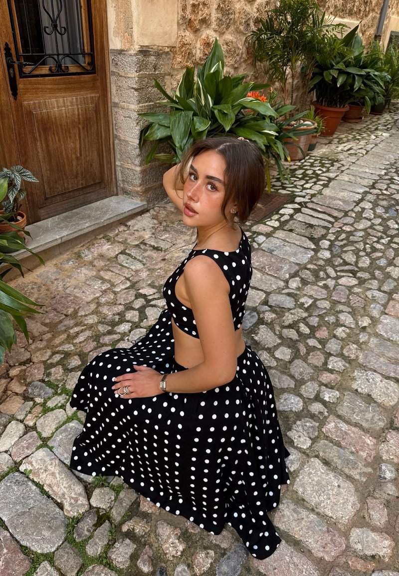 Jeune femme en robe blanche à pois noirs assise sur une rue pavée près d’une porte et de plantes dans des pots en terre cuite.