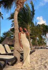 Strick-Maxikleid in Off-White mit einem Diamantmuster, V-Ausschnitt und dünnen Trägern. Der Hintergrund zeigt Palmen und Strandstühle.