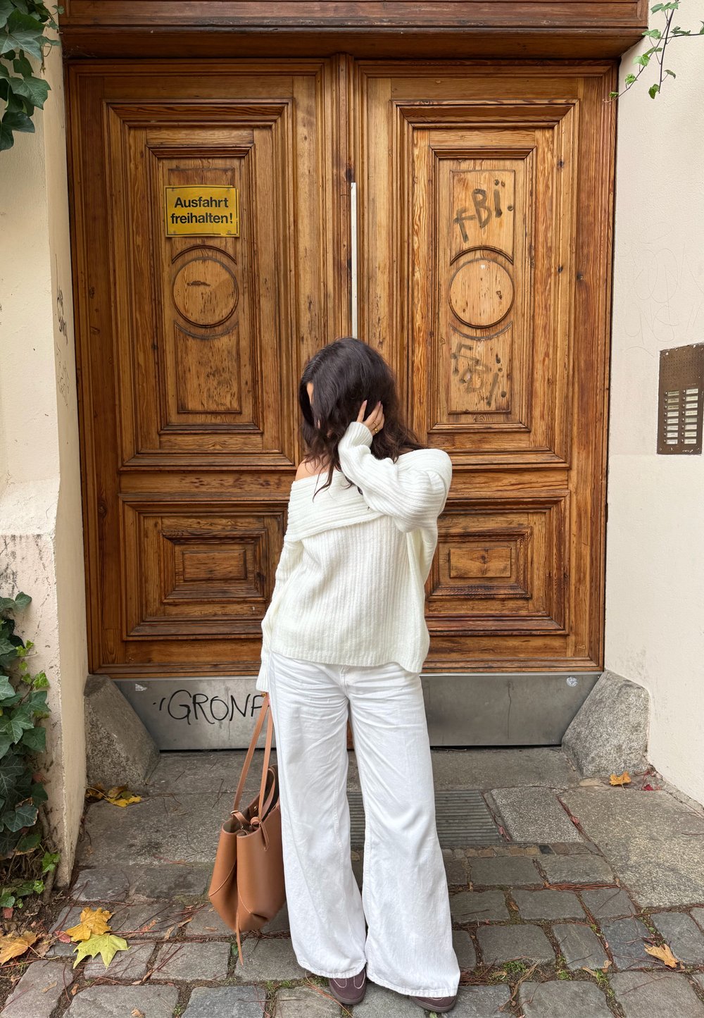 Világos krémszínű bordázott pulóver, széles fehér nadrág, barna bőr táska, faragott mintás és graffitivel díszített faajtók előtt állva.