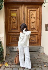 Világos krémszínű bordázott pulóver, széles fehér nadrág, barna bőr táska, faragott mintás és graffitivel díszített faajtók előtt állva.