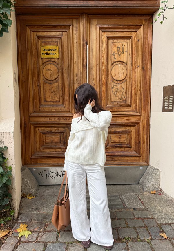 Hele kreemikas soonikkoes kampsun, laiad valged püksid, pruun nahast kott, seisab puidust ukse ees, millel on peen mustrite ja graffitiga kaunistus.