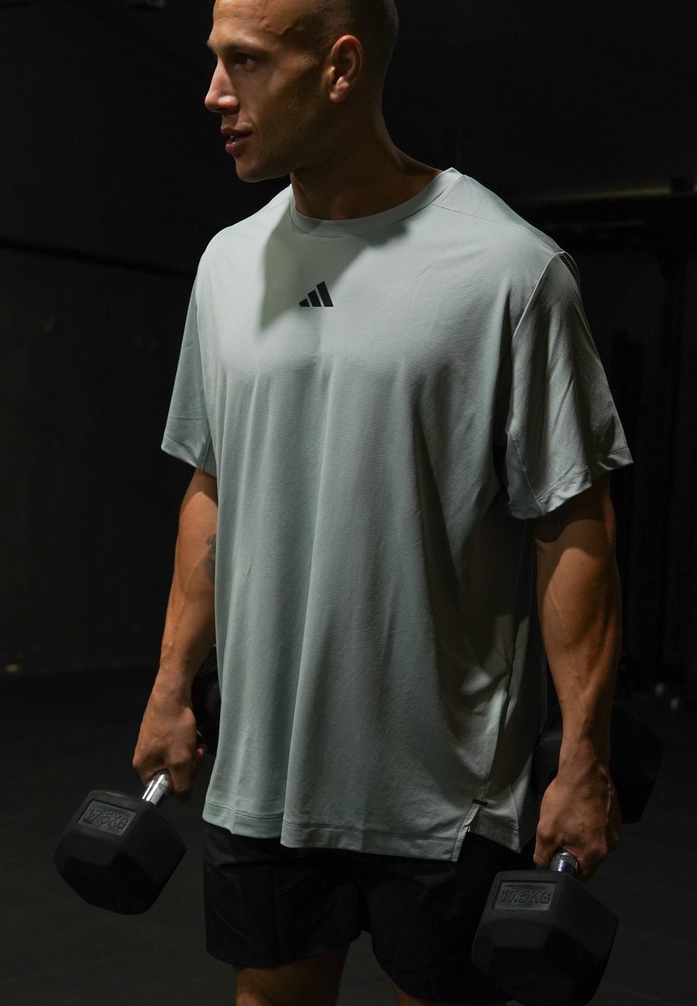 Homme en chemise Adidas gris clair et short noir tenant des haltères de 17,5 kg dans chaque main, debout dans une salle de sport peu éclairée.