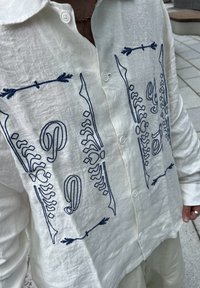 Linne skjorta i off-white med blå broderade mönster med skelettmotiv, dekorativa linjer och knappar fram.
