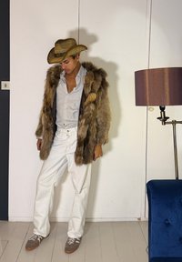 Άνδρας με καουμπόικο καπέλο, παλτό από γούνα, ριγέ πουκάμισο, άσπρο παντελόνι και αθλητικά παπούτσια στέκεται μέσα σε δωμάτιο κοντά σε επιδαπέδιο φωτιστικό και μπλε καναπέ.