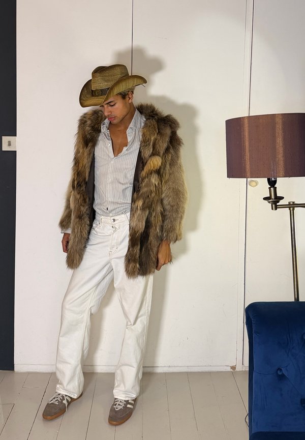 Άνδρας με καουμπόικο καπέλο, παλτό από γούνα, ριγέ πουκάμισο, άσπρο παντελόνι και αθλητικά παπούτσια στέκεται μέσα σε δωμάτιο κοντά σε επιδαπέδιο φωτιστικό και μπλε καναπέ.