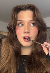 Osoba o brązowych włosach trzyma brązową kredkę do ust blisko ust, nosi złoty pierścień w przegrodzie nosa i naszyjnik z krzyżem; skóra ma jasne piegi.