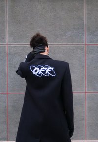 Personne aux cheveux bouclés portant un manteau noir avec le texte blanc « OFF » au dos, des gants noirs, debout contre un mur carrelé gris.