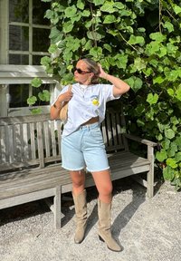 Witte cropped t-shirt met gele graphic, lichtblauwe denim shorts, tan suède kniehoge laarzen, en een geweven tas tegen een groene achtergrond.